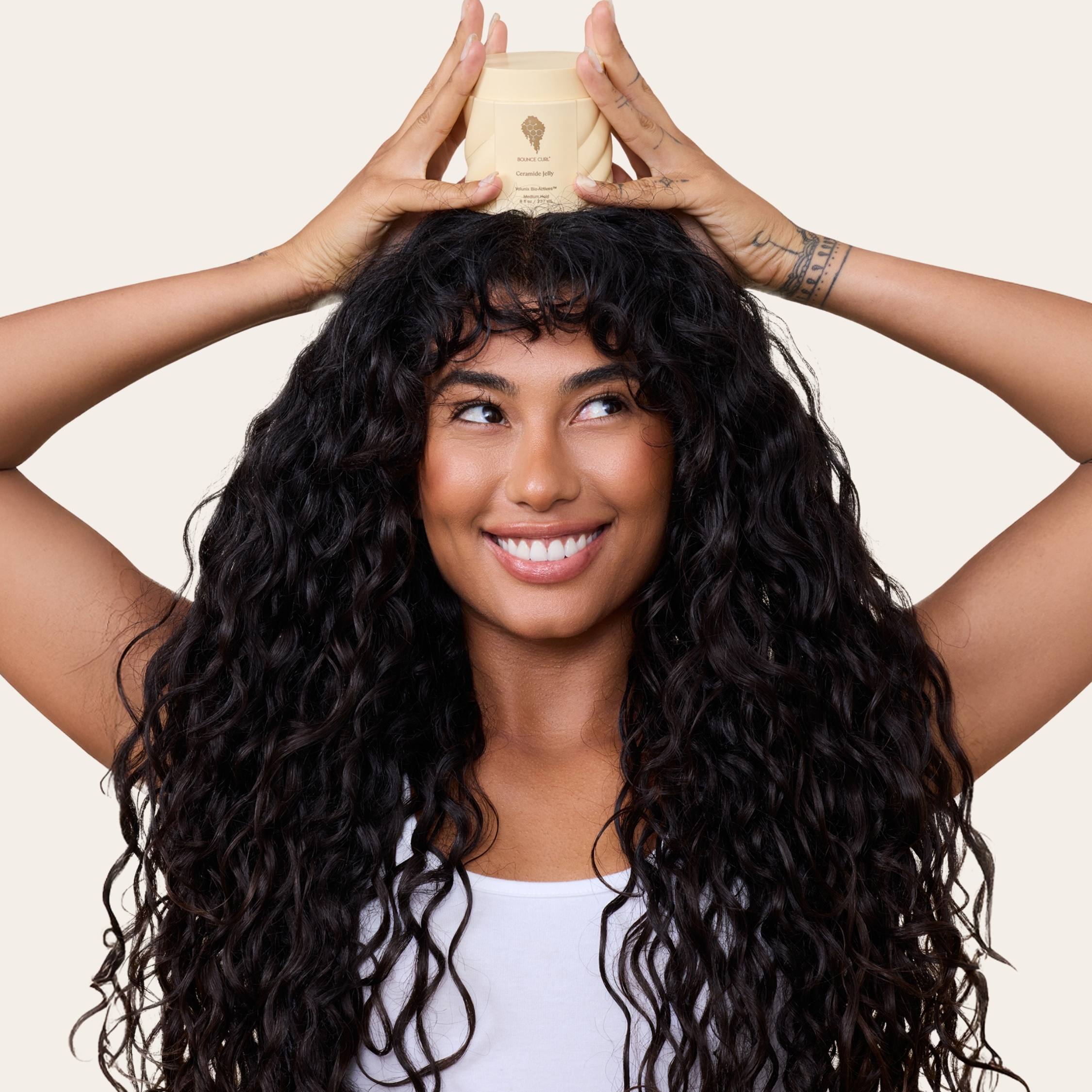 Woman with long, wavy hair holding a product container above her head against a beige background